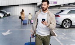Bărbat cu troler și telefon mobil într-o parcare de aeroport, pregătit să preia o mașină închiriată.