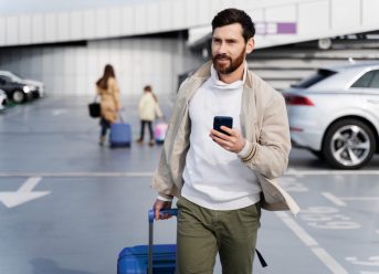 Bărbat cu troler și telefon mobil într-o parcare de aeroport, pregătit să preia o mașină închiriată.