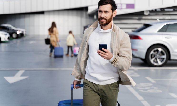 Bărbat cu troler și telefon mobil într-o parcare de aeroport, pregătit să preia o mașină închiriată.